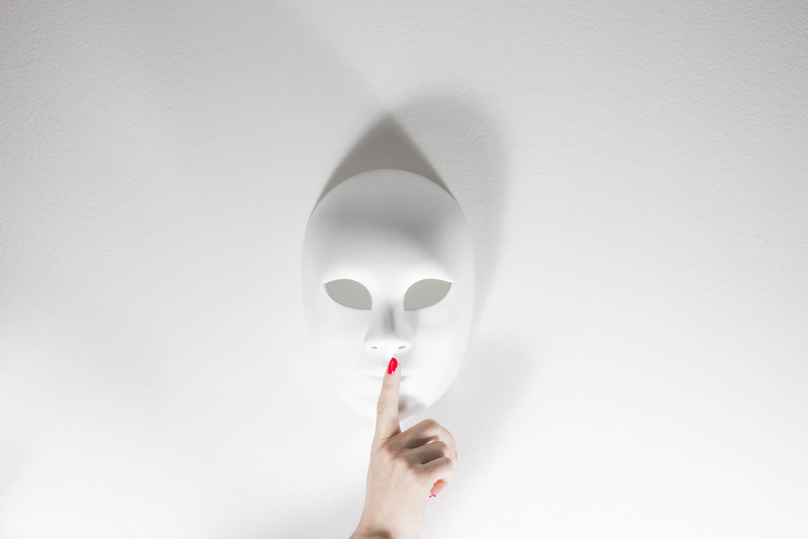 Un masque blanc avec une main humaine faisant un geste de silence sur un fond blanc lumineux, créant une atmosphère myst